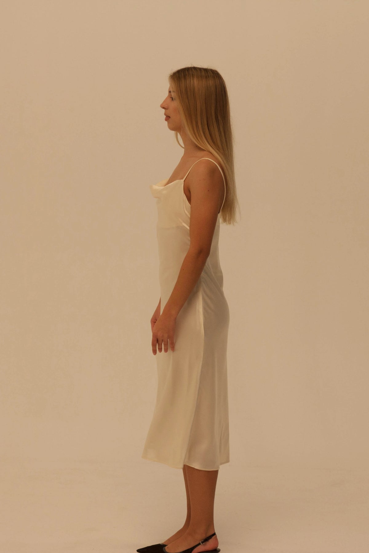 A woman in profile wearing a champagne-colored spaghetti strap midi slip dress, standing against a plain beige studio background.