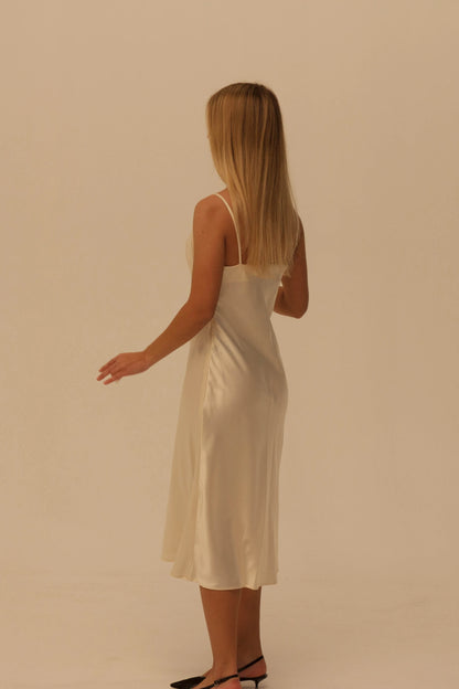A woman with long blonde hair is shown from the back wearing a champagne silk slip dress and black pointed-toe heels against a plain beige studio background.
