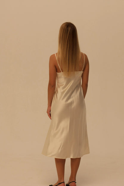 A woman with long blonde hair is shown from behind wearing a champagne silk slip dress with spaghetti straps, standing against a solid beige studio background.