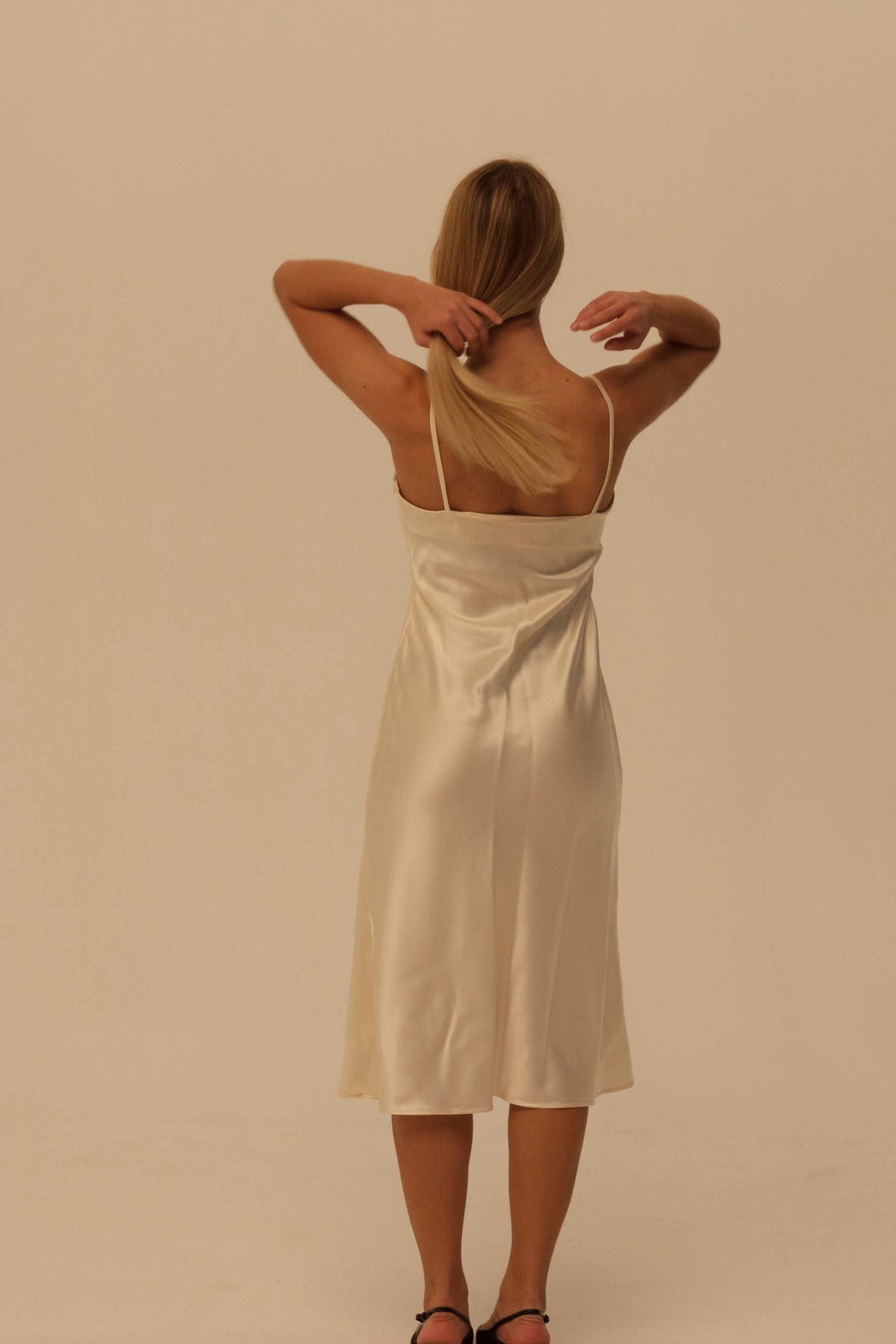 A woman viewed from behind wearing a cream silk slip dress with spaghetti straps, standing against a solid beige studio background.