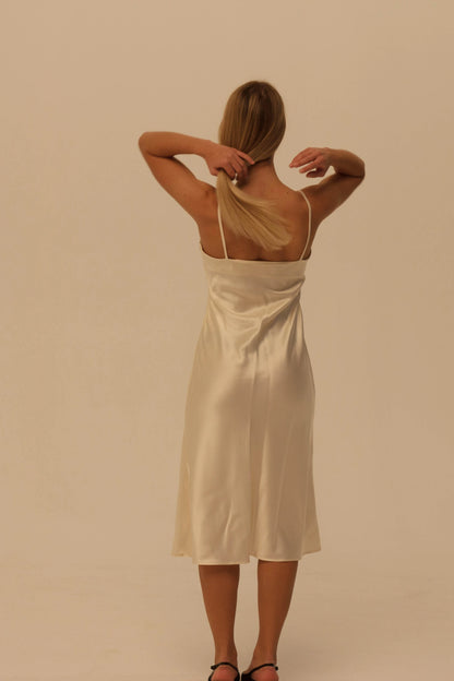 A woman viewed from behind wearing a cream silk slip dress with spaghetti straps, standing against a solid beige studio background.