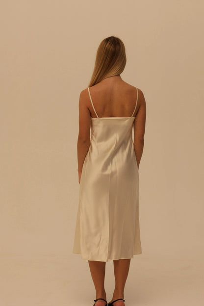 A woman viewed from behind wearing a champagne colored silk slip dress with thin spaghetti straps and a low back, standing against a solid warm beige studio background.