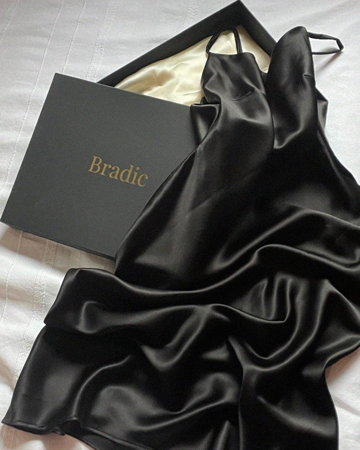 Black bias cut silk slip dress laying flat across Bradic packaging.