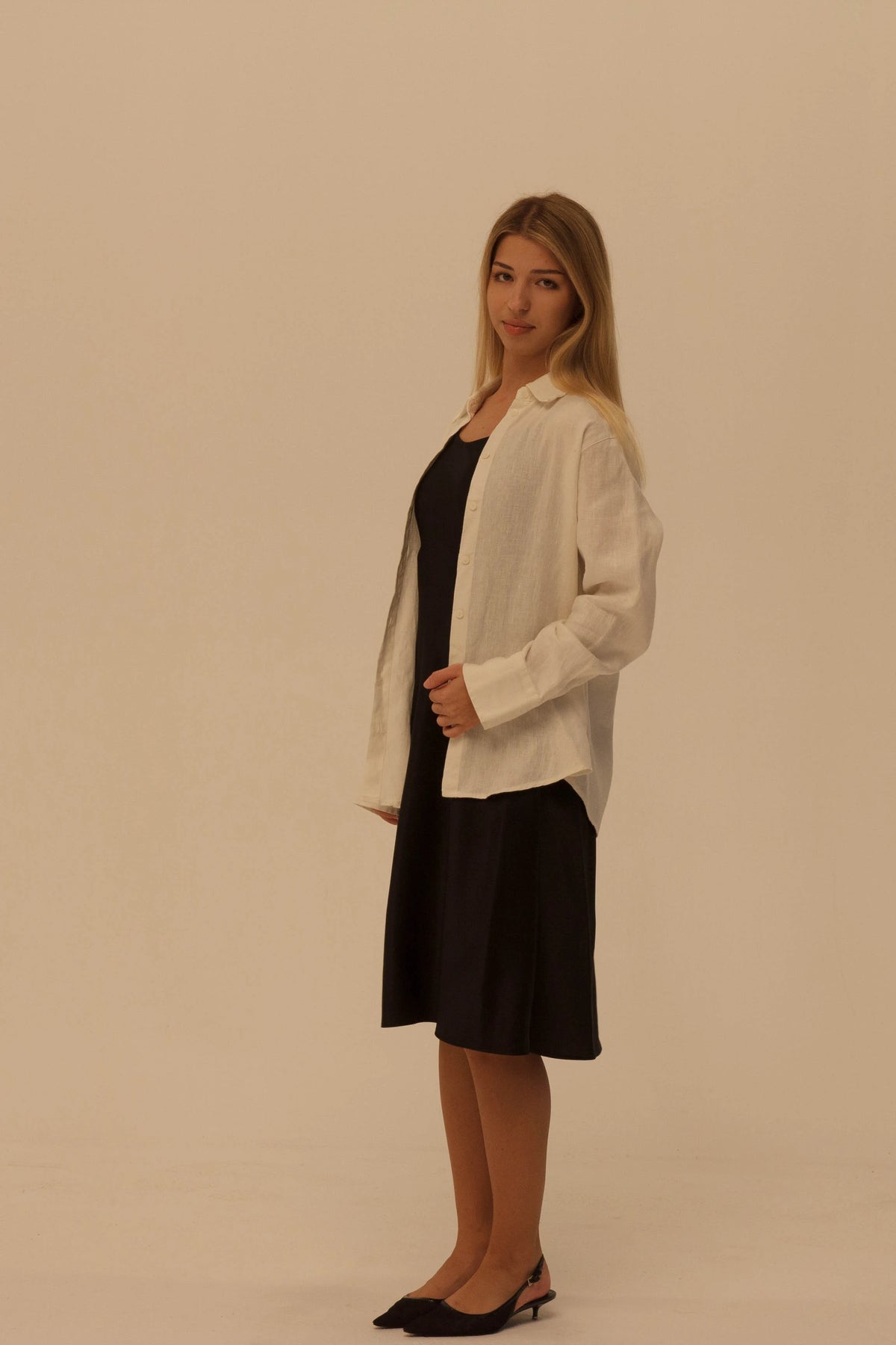 A woman modeling a white long-sleeve linen-blend shirt jacket over a black silk slip dress, standing against a plain beige studio background.