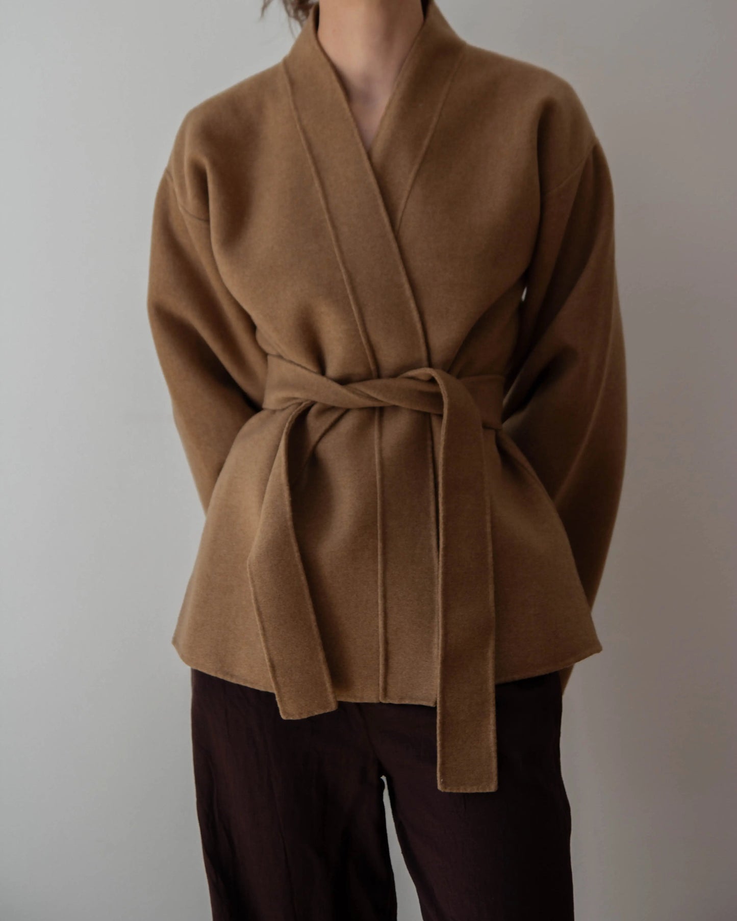 A woman wearing a camel brown belted cashmere wrap coat with wide sleeves, paired with dark brown pants, against a plain light gray background.