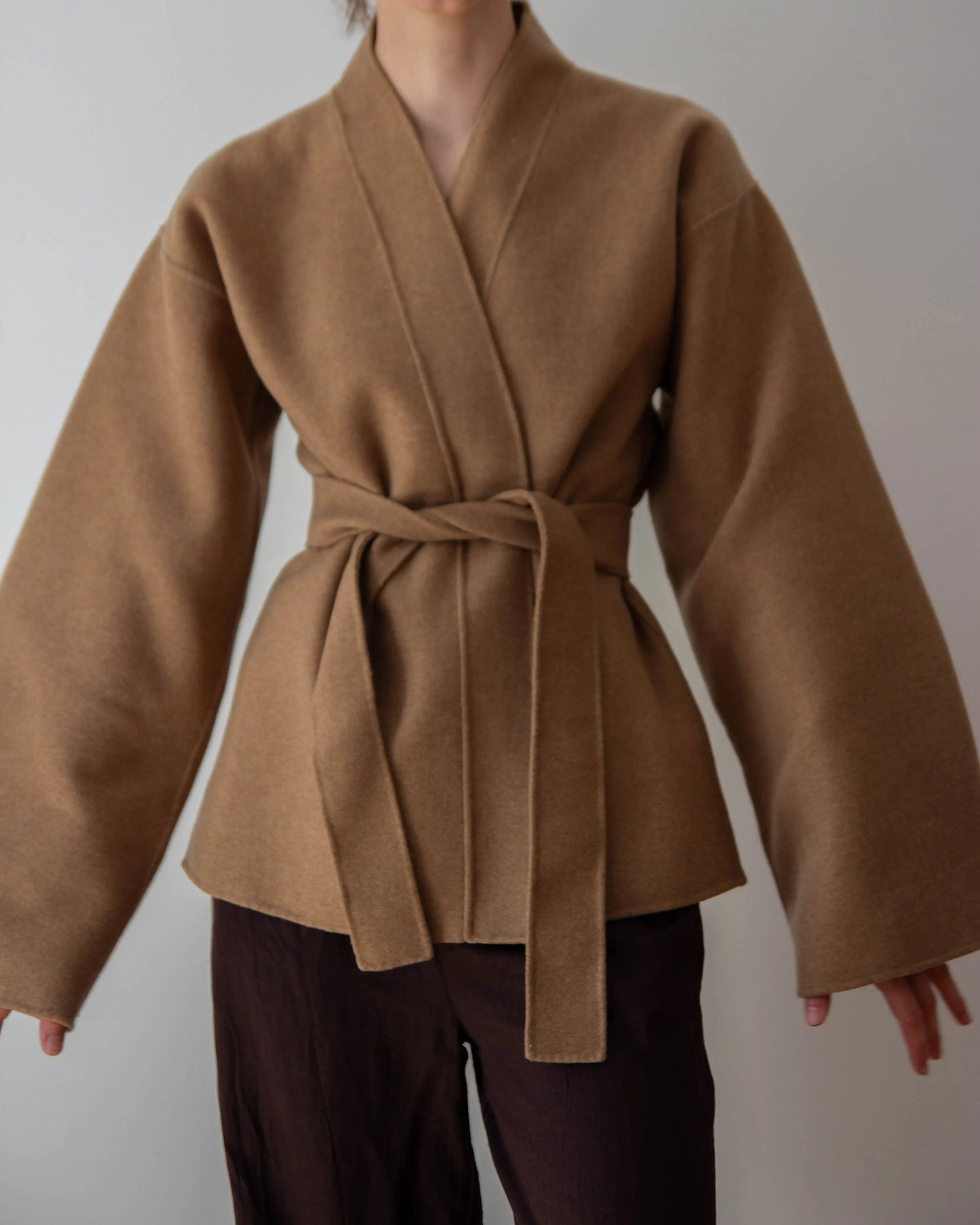 A woman wearing a brown belted double faced cashmere wrap coat with wide sleeves, paired with dark brown pants, against a plain light gray background.