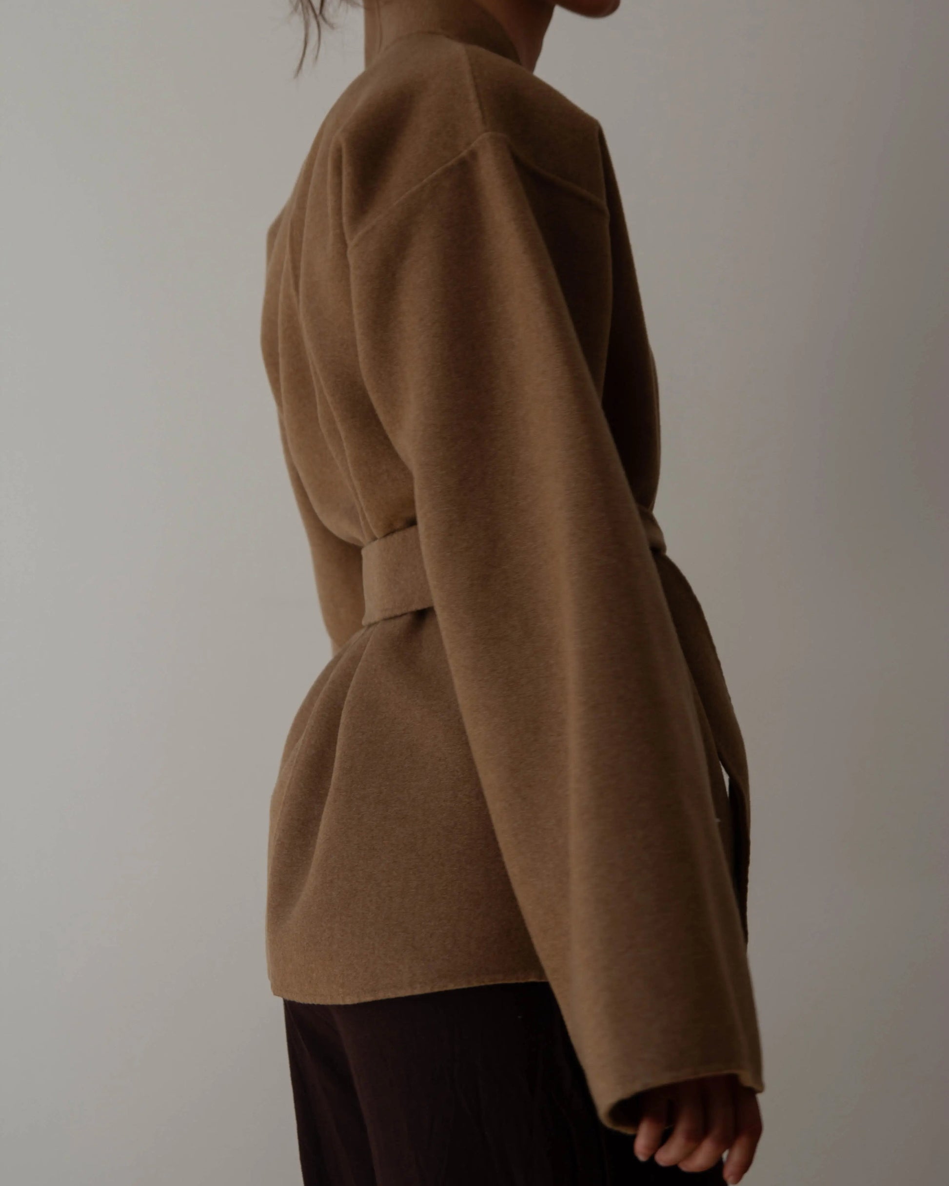 A person wearing a camel brown belted cashmere wrap coat with wide sleeves, paired with dark brown pants, against a plain light gray background.
