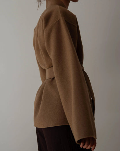 A person wearing a camel brown belted cashmere wrap coat with wide sleeves, paired with dark brown pants, against a plain light gray background.