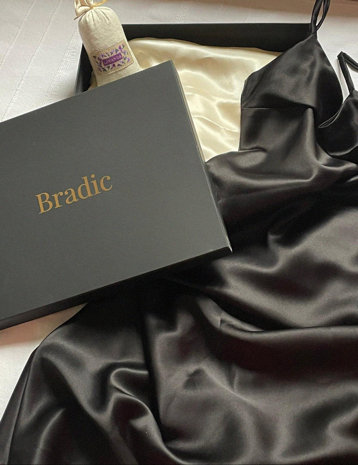 Black bias cut silk slip dress flat lay across Bradic packaging.
